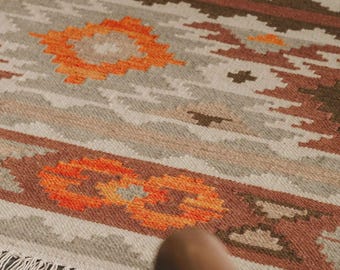 May include: A close-up of a woven rug with a geometric pattern in shades of orange, brown, and beige. The rug has a fringe border.