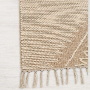 Peut inclure: Un tapis tiss&eacute; beige et blanc avec un motif g&eacute;om&eacute;trique et des franges sur le bord inf&eacute;rieur.