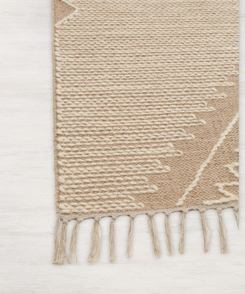 Peut inclure: Un tapis tiss&eacute; beige et blanc avec un motif g&eacute;om&eacute;trique et des franges sur le bord inf&eacute;rieur.