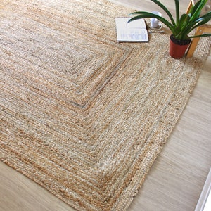 A natural fiber rug with a light brown color and a woven texture. The rug has a border that is slightly darker than the rest of the rug. The rug is on a light wood floor.