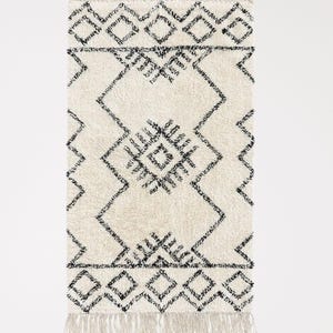 May include: A white and black geometric patterned rug with fringe on all sides. The rug has a diamond pattern with black lines.