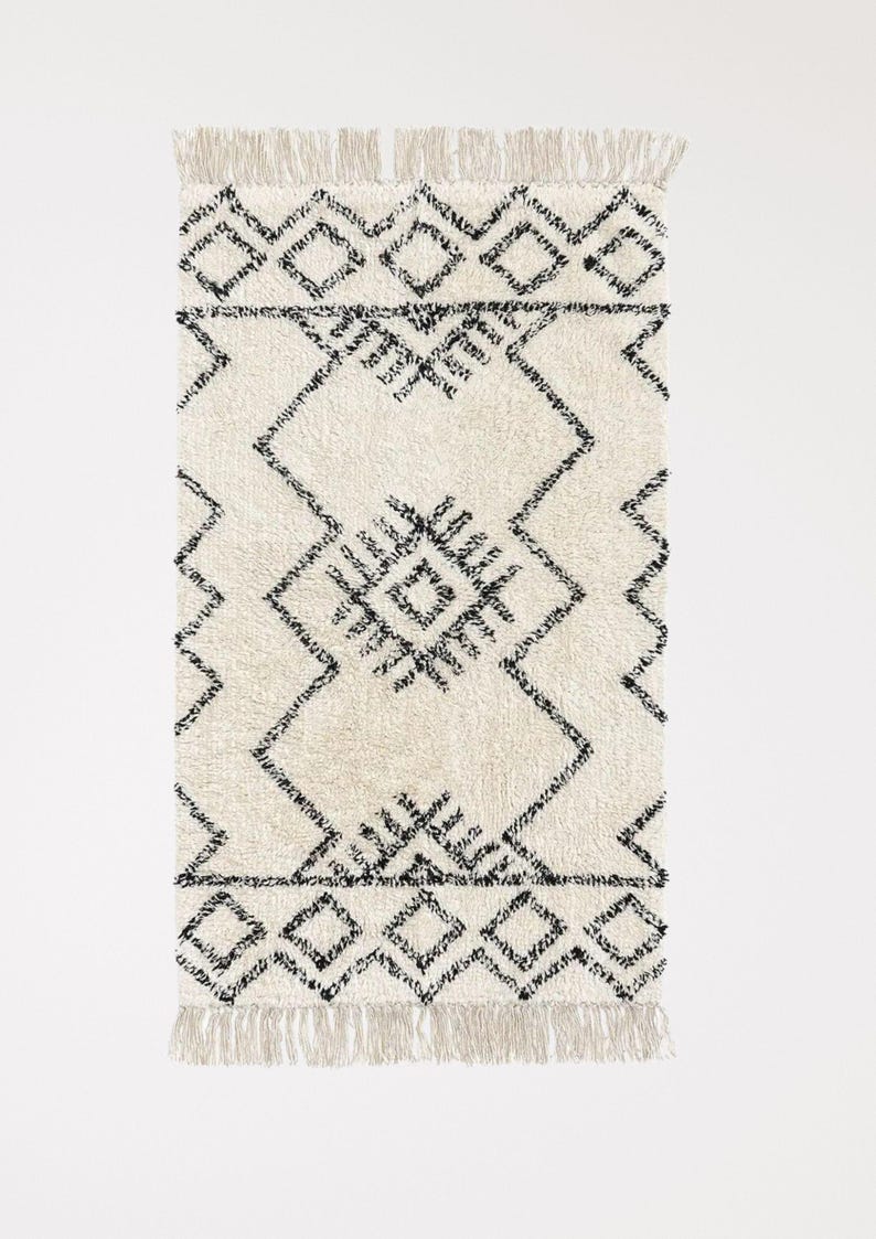 May include: A white and black geometric patterned rug with fringe on all sides. The rug has a diamond pattern with black lines.