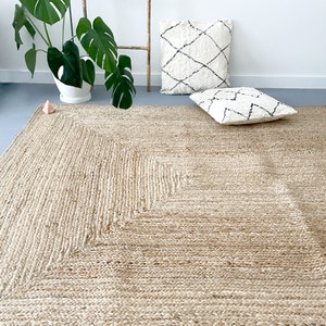 A square, natural fiber rug with a diamond pattern in the center. The rug is a light beige color and is made of woven jute. Two white throw pillows with black geometric patterns are placed on the rug.