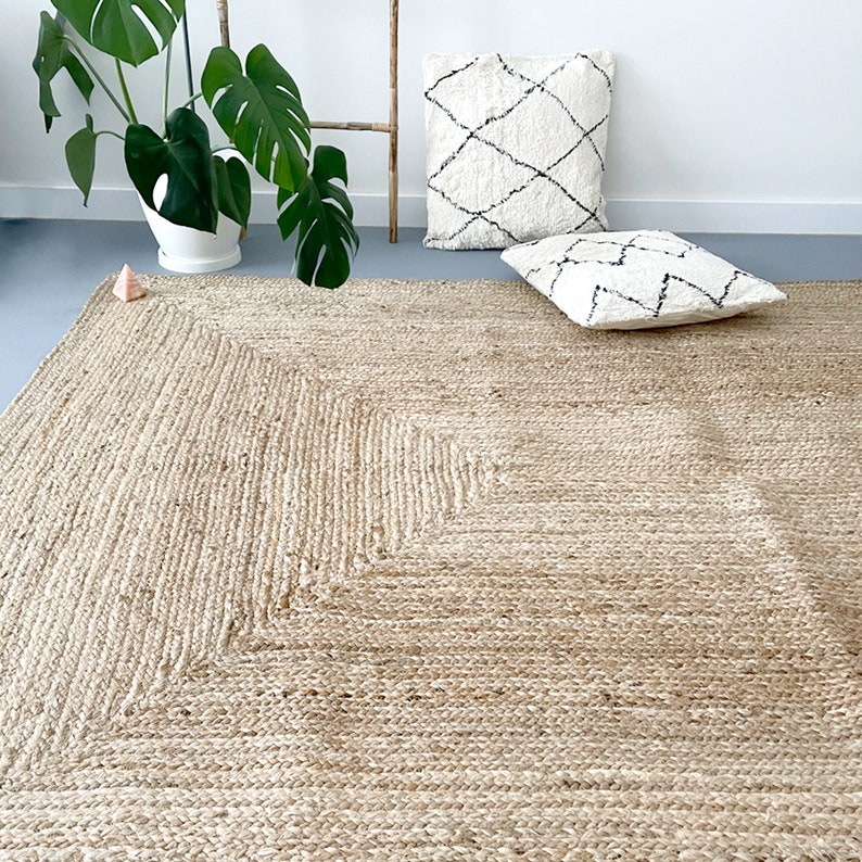 A square, natural fiber rug with a diamond pattern in the center. The rug is a light beige color and is made of woven jute. Two white throw pillows with black geometric patterns are placed on the rug.