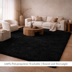 May include: A large, rectangular black rug dominates the foreground of a living room scene. The rug is made of a plush material. The room features a white sofa, a wooden coffee table, and a cream-colored armchair. The text at the bottom reads: "100% Polypropylene Washable | Round and Rectangle."