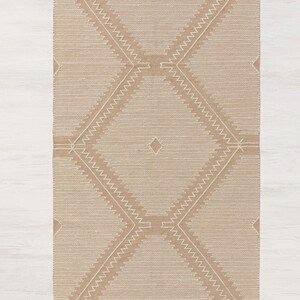 Peut inclure: Un tapis beige avec un motif g&eacute;om&eacute;trique blanc. Le tapis a une bordure en franges.