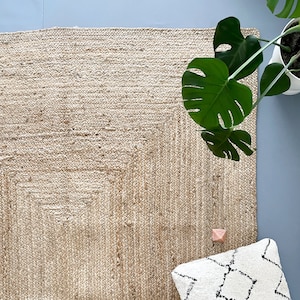 A natural fiber jute rug with a square pattern. The rug is beige and has a light brown border. A white pillow with black diamond patterns is on the rug. A green plant is in the upper right corner of the image.
