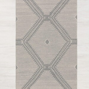Peut inclure: Un tapis gris et blanc &agrave; motifs g&eacute;om&eacute;triques avec des franges sur tous les c&ocirc;t&eacute;s. Le tapis a un motif en forme de diamant avec un plus petit diamant au centre.