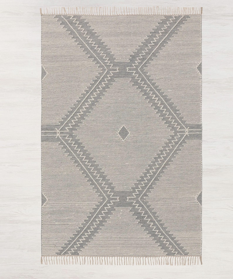 Peut inclure: Un tapis gris et blanc &agrave; motifs g&eacute;om&eacute;triques avec des franges sur tous les c&ocirc;t&eacute;s. Le tapis a un motif en forme de diamant avec un plus petit diamant au centre.