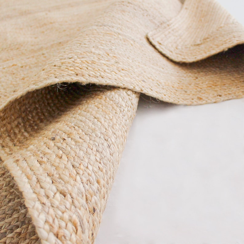 A close-up of a natural fiber rug made of woven jute. The rug is a light beige color and has a rough texture. The rug is folded in half, showing the underside of the rug.