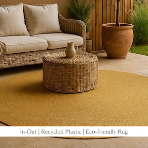 May include: A round, mustard-yellow outdoor rug made of recycled plastic. A woven wicker coffee table and a beige pitcher sit on top of the rug. A wicker sofa with beige cushions is in the background.