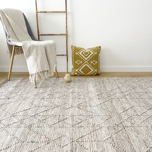 May include: A light brown woven rug with a diamond pattern. The rug is on a wooden floor in a room with a white wall. A chair with a white throw blanket and a yellow pillow with a white geometric design are in the background.