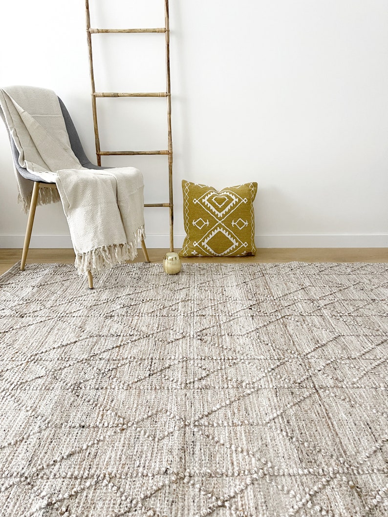 May include: A light brown woven rug with a diamond pattern. The rug is on a wooden floor in a room with a white wall. A chair with a white throw blanket and a yellow pillow with a white geometric design are in the background.