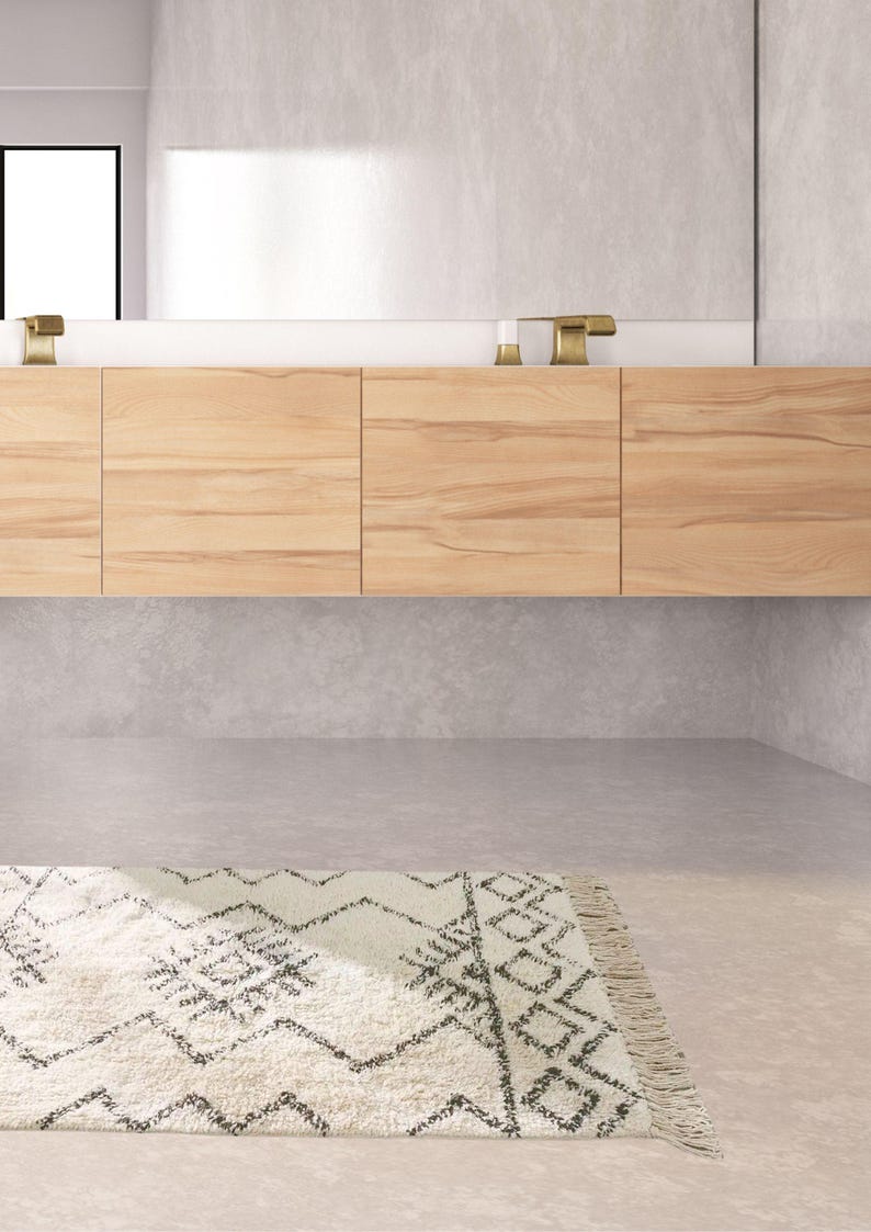 May include: A white bathroom vanity with a wooden cabinet and a white countertop. The vanity has two gold faucets and a white rug with a black geometric pattern is on the floor.