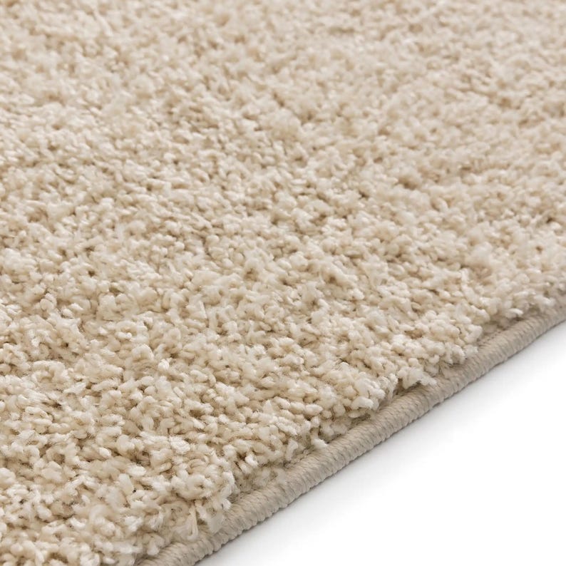 May include: Close-up of a beige, plush rug with a soft, textured surface. The rug has a thick pile, creating a cozy and inviting appearance. The edges are neatly finished with a subtle border.