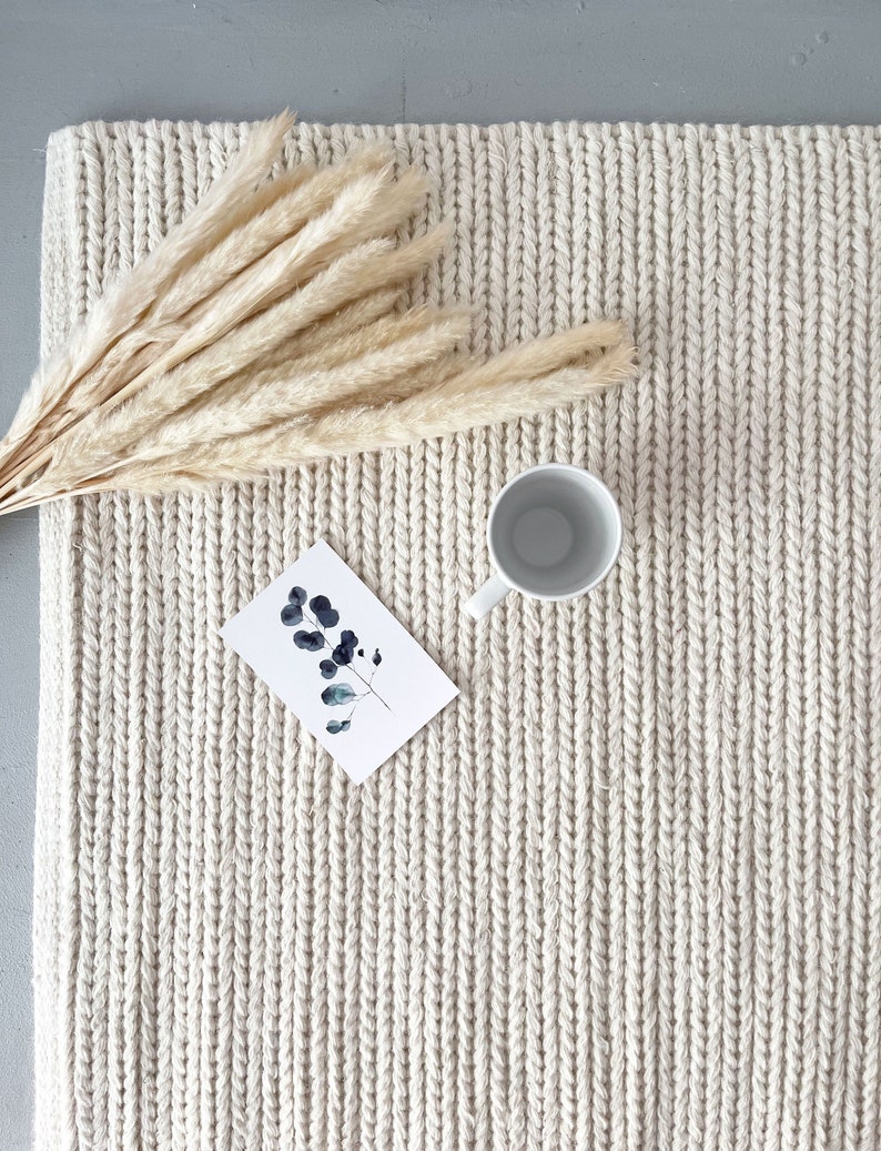 May include: A cream-colored woven rug with a textured surface. The rug is decorated with a bouquet of dried pampas grass and a white ceramic cup.