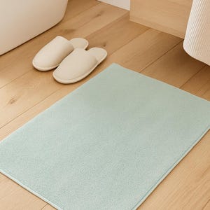 Op de afbeelding: Een lichtblauw rechthoekig badmat met een witte rand ligt op een houten vloer. Twee crèmekleurige slippers zijn in de buurt geplaatst. De scène suggereert een badkameromgeving, met de nadruk op comfort en netheid.