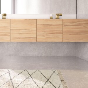 May include: A white bathroom rug with a black diamond pattern. The rug has a fringe border and is placed in front of a wooden cabinet with a white countertop.