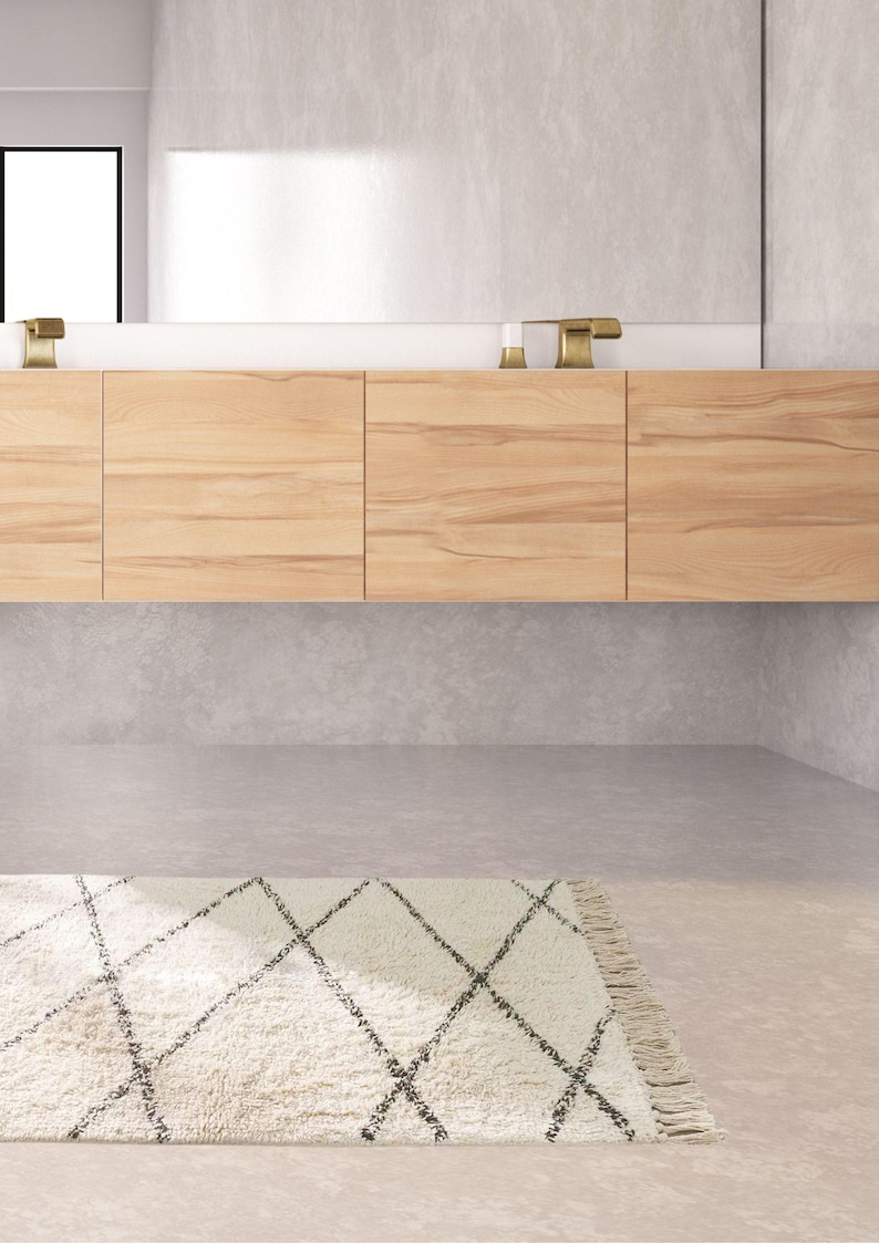 May include: A white bathroom rug with a black diamond pattern. The rug has a fringe border and is placed in front of a wooden cabinet with a white countertop.