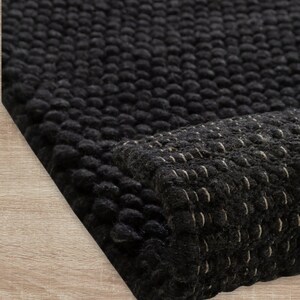 Peut inclure: Gros plan sur un tapis noir texturé avec une surface à nœuds en relief. Le bord du tapis est replié, révélant un dessous contrastant avec des coutures blanches. Le tapis est posé sur une surface en bois clair.