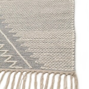 Peut inclure: Un tapis tiss&eacute; gris et blanc avec un motif g&eacute;om&eacute;trique et des franges sur le bord inf&eacute;rieur.