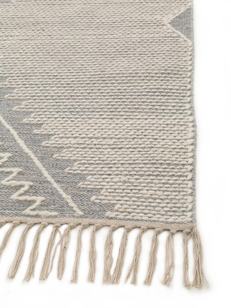 Peut inclure: Un tapis tiss&eacute; gris et blanc avec un motif g&eacute;om&eacute;trique et des franges sur le bord inf&eacute;rieur.
