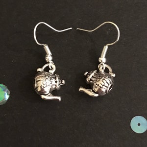 Peut inclure: Une paire de boucles d'oreilles en forme de théière argentées. Les boucles d'oreilles sont petites et détaillées, avec un motif de théière. Les boucles d'oreilles sont sur un fond noir.
