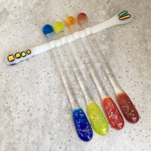 May include: Set of seven glass stirring sticks with colourful, iridescent tips. The tips are blue, yellow, orange, red, and two shades of orange. The sticks are clear glass and have a white base.