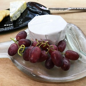 May include: A clear glass serving tray with a round, white cheese wheel, a bunch of red grapes, and a silver knife. The tray has a unique, organic shape.