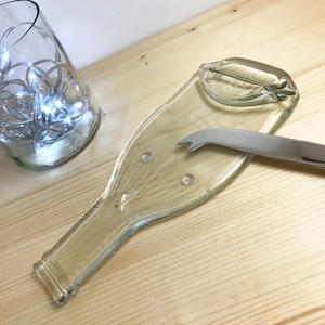 May include: A clear glass bottle shaped like a wine bottle, repurposed as a serving tray for cheese and crackers. A silver cheese knife is resting on the tray.