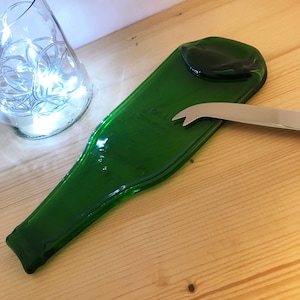 May include: A green glass bottle repurposed into a serving tray with a cheese knife resting on top. The bottle has a smooth, rounded shape and is perfect for serving cheese, crackers, or other appetizers.
