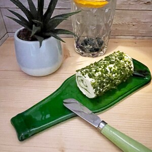May include: A green glass bottle shaped serving tray with a roll of herb and cream cheese and a butter knife.