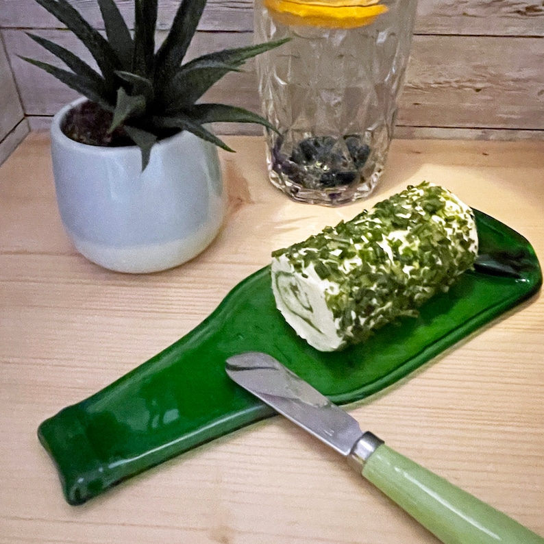 May include: A green glass bottle shaped serving tray with a roll of herb and cream cheese and a butter knife.