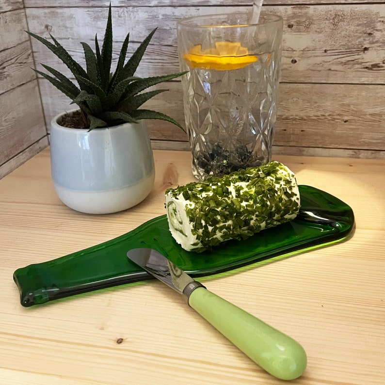 May include: A green glass bottle shaped serving tray with a roll of herb-infused cheese and a green handled butter knife.