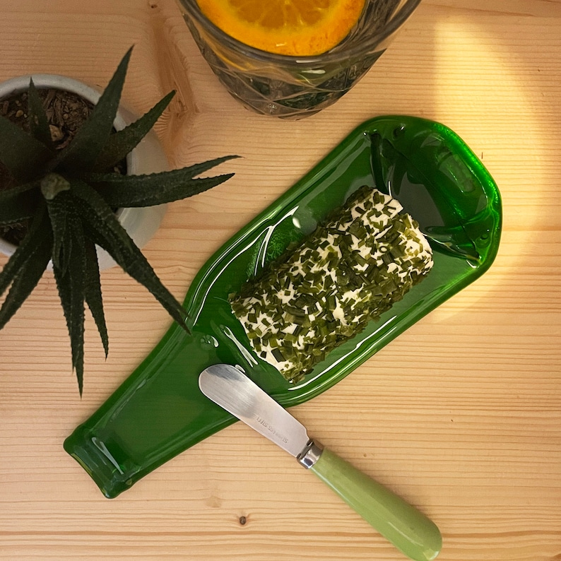May include: A green glass bottle shaped serving dish with a roll of herb-infused cheese and a green handled butter knife.