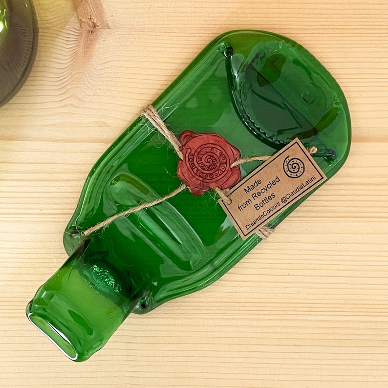 May include: A green glass serving dish made from recycled bottles. The dish is shaped like a bottle and has a small lip around the edge. It is tied with twine and has a wax seal with a logo that says "DreaminColours". The tag attached to the dish reads "Made from Recycled Bottles DreaminColours @ClaudiaLatini"
