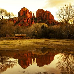 May include: A scenic view of red rock formations reflected in a puddle of water. The rocks are tall and jagged, and the water is still and clear. The sky is a soft orange and yellow, and the trees are bare and brown.