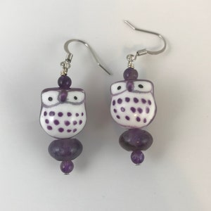 May include: A pair of dangle earrings featuring a white owl charm with purple polka dots, a large purple bead, and a smaller purple bead at the bottom. The earrings have silver hooks.