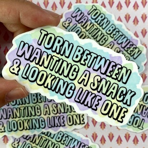 May include: A sticker with a colourful, watercolour-like background featuring the text "Torn between wanting a snack & looking like one."