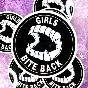 Può includere: Un gruppo di adesivi in bianco e nero con un design circolare. Gli adesivi presentano un set di zanne stilizzate e la scritta "GIRLS BITE BACK" in grassetto. Lo sfondo è una superficie viola strutturata.