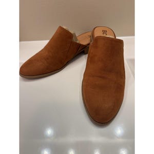 May include: A pair of brown suede slip-on mules. The shoes have a pointed toe and a low heel. The interior of the shoe is visible. The shoes are on a white surface.