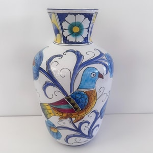 Op de afbeelding: Een witte keramische vaas met een handgeschilderd ontwerp. De vaas heeft een blauwe vogel met oranje en gele accenten, omringd door blauwe bloempatronen. De rand en de basis hebben blauwe en gele bloemendetails.