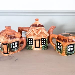 Peut inclure: Service à thé en céramique en forme de maison de campagne, comprenant une théière, un sucrier et une crémière. L'ensemble présente un toit de chaume, avec des accents marron, orange et vert. Les fenêtres et portes sont peintes en noir.