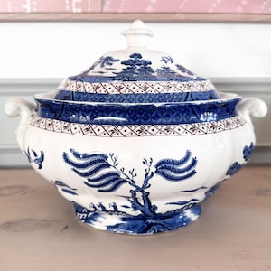 Op de afbeelding: Een witte en blauwe keramische terrine met deksel. De terrine heeft een klassiek Willow-patroon met een blauw-witte kleurstelling. Het ontwerp omvat een landschap met bomen en een brug. De terrine heeft twee handvatten en een decoratieve knop.