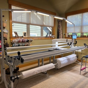 May include: A white quilting frame with attached lighting, a rolled-up quilt, and a black chair. The frame is in a well-lit room with windows and a view of the outdoors. Quilts hang on the wall in the background.