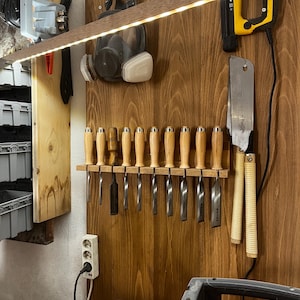 Peut inclure: Un mur en bois avec une variété d'outils accrochés. Il y a des ciseaux, une scie, un respirateur et un outil électrique. Le mur est éclairé par une bande de lumières LED.