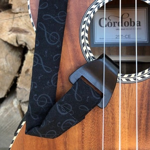 Może przedstawiać: Brązowy drewniany ukulele z czarno-białym wzorzystym otworem rezonansowym. Ukulele ma czarny pasek z wzorem nut muzycznych. Ukulele jest oznaczone "Cordoba" i "25T-CE".