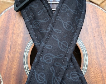 May include: A black ukulele strap with a treble clef pattern. The strap is attached to a ukulele with a wood body and a white fretboard.
