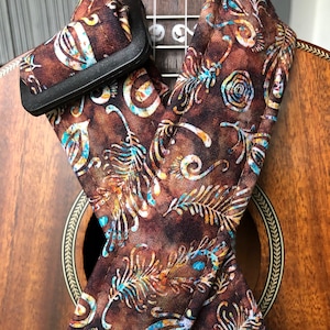 May include: A brown ukulele strap featuring a pattern of stylized leaves and swirls in blue, yellow, and white. The strap is draped over a wooden ukulele, with a black plastic buckle.