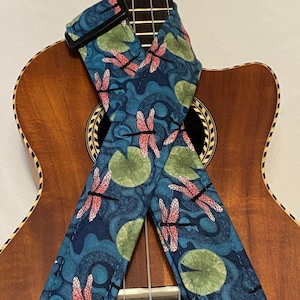 Puede incluir: Una correa de ukelele azul con un patrón de libélula y nenúfar. La correa se muestra en un ukelele.